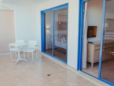 Moderní balkon s bílým nábytkem před modrými skleněnými posuvnými dveřmi hotelového pokoje.