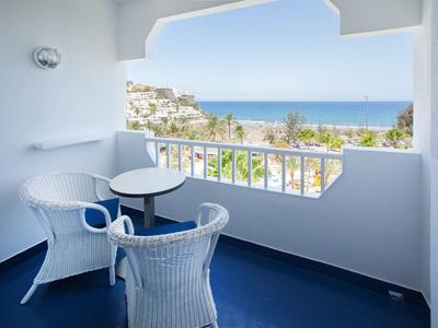 Balkon mit weißen Korbstühlen und Tisch, blauer Boden, Blick auf Meer und Häuser am Hang.