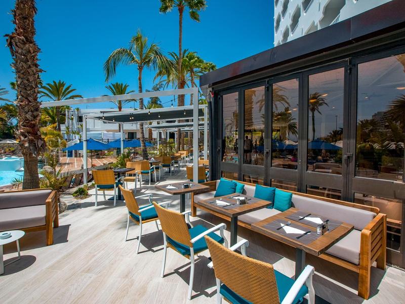 Terrazza esterna dell'hotel con tavoli e sedie vicino alla piscina sotto palme in una giornata di sole.