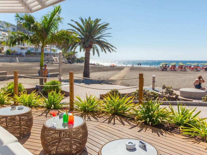 Terrazza sulla spiaggia con tavoli in vimini, palme e spiaggia di sabbia sotto un cielo limpido.