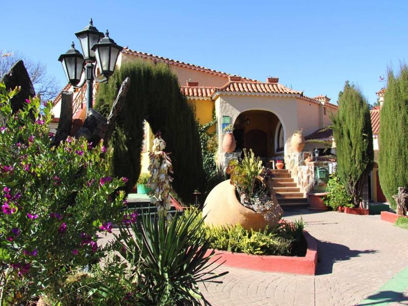 Haus mit rotem Ziegeldach, Säulenportikus, grünem Garten und bunten Blumen bei klarem Himmel.