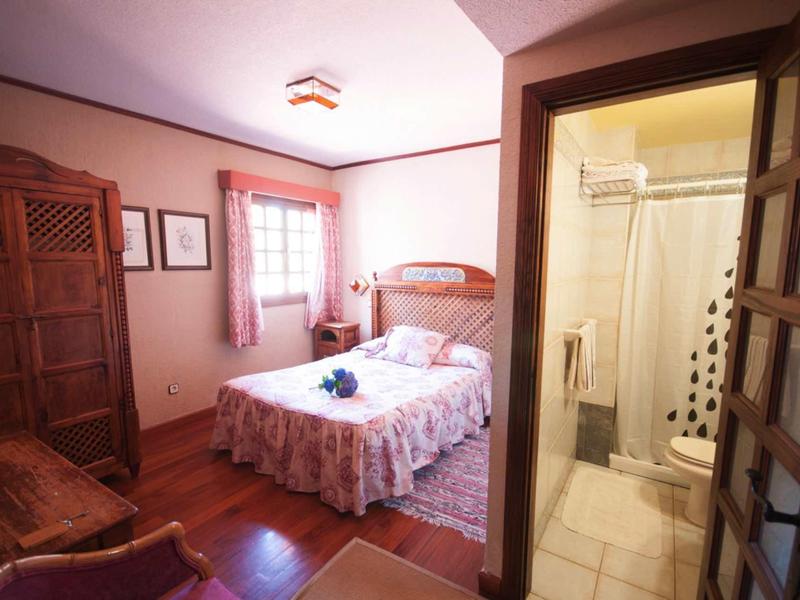 Gemütliches Schlafzimmer mit Holzschrank, Doppelbett, rosa Vorhängen und angrenzendem Bad mit Dusche.