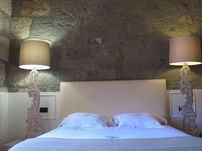 Letto matrimoniale semplice con lenzuola bianche davanti a un muro di pietra, affiancato da due lampade decorative.