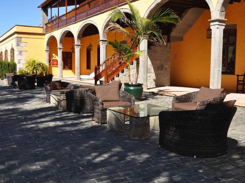 Area salotto con mobili in vimini e tavoli davanti a un edificio giallo con archi e balcone.