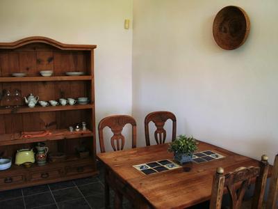 Coin repas rustique avec table en bois, chaises et étagère remplie de vaisselle contre un mur blanc.