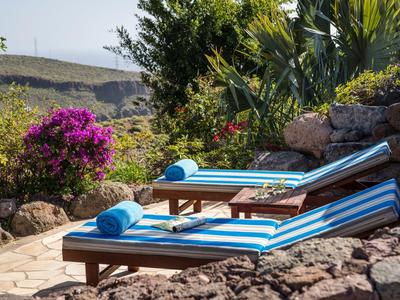 Due lettini con cuscini blu e bianchi e asciugamani accanto a un giardino con piante e montagne sullo sfondo.