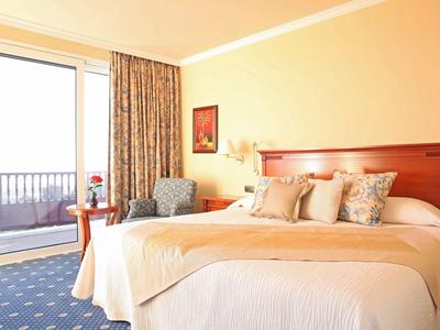 Camera d'hotel luminosa con un grande letto, poltrone e balcone con vista.