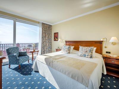 Camera d'hotel luminosa con un grande letto, due poltrone e un balcone con vista sul mare.