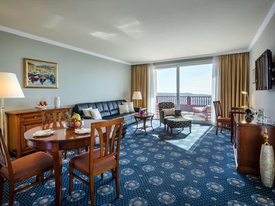 Camera d'hotel luminosa con moquette blu, mobili in legno e balcone con vista sul mare.