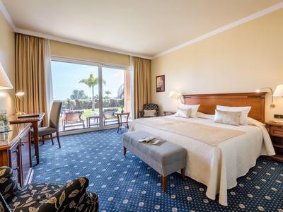 Camera d'hotel luminosa con letto matrimoniale, scrivania, poltrone e balcone con vista sulle palme.