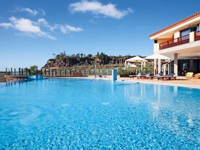 Grande piscina all'aperto con acqua azzurra cristallina accanto a un edificio alberghiero moderno e ombrelloni.
