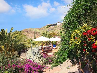 Vista di una terrazza con ombrellone in un giardino rigoglioso e colline sullo sfondo.