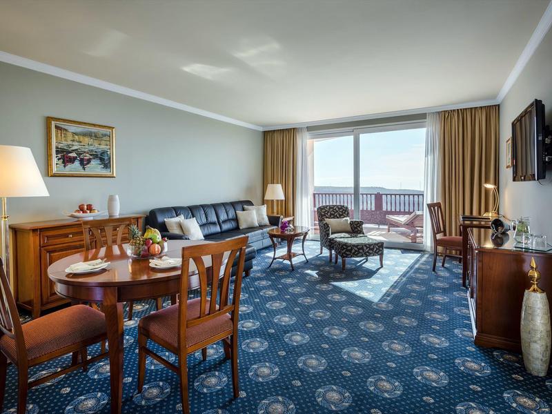 Camera d'hotel luminosa con moquette blu, mobili in legno e balcone con vista sul mare.