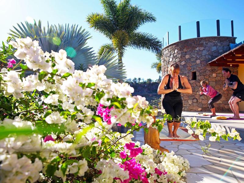 Due donne che praticano yoga su una terrazza con piante tropicali e palme sullo sfondo.
