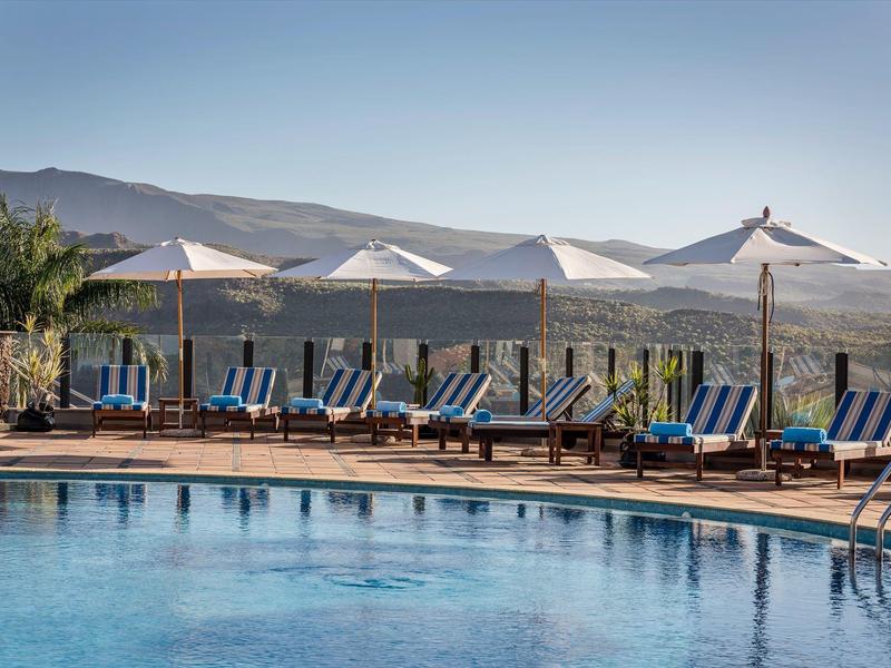 Area piscina con lettini e ombrelloni davanti a uno scenario montano sotto un cielo sereno.