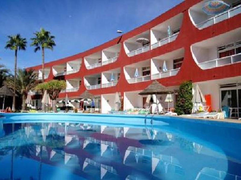 Hôtel avec balcons rouges et piscine bleue par une journée ensoleillée, entouré de palmiers.