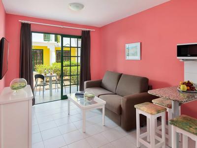 Helles Wohnzimmer mit rosa Wand, grauem Sofa, weißem Tisch, Fernseher und Balkon mit Gartenblick.