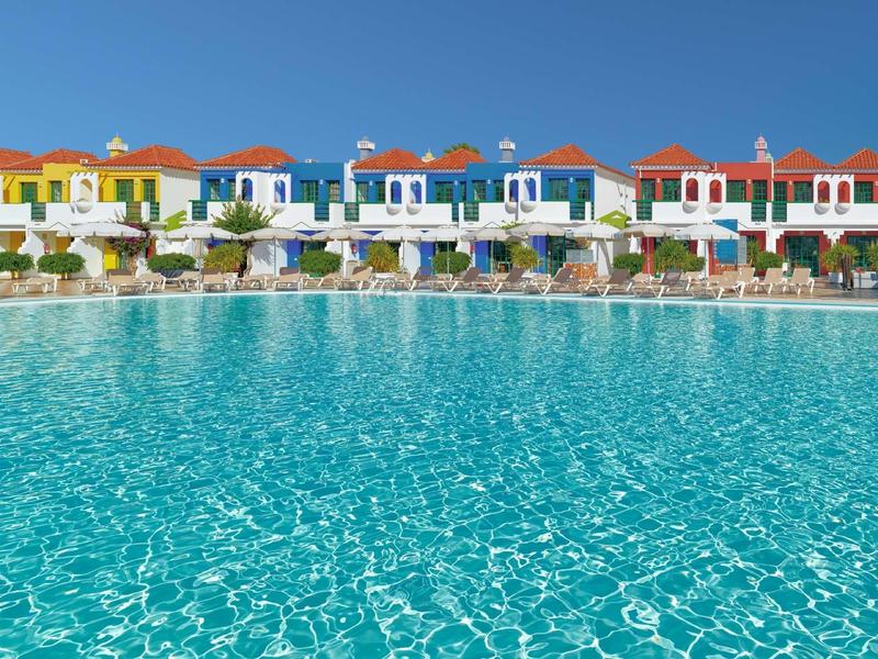 Reihe bunter Häuser am großen, klaren Schwimmbad unter blauem Himmel.