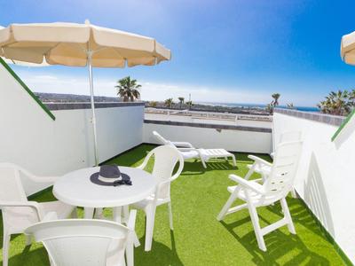Dachterrasse mit grünem Boden, weißen Stühlen, Tisch, Sonnenschirmen und Blick auf Himmel und Meer.