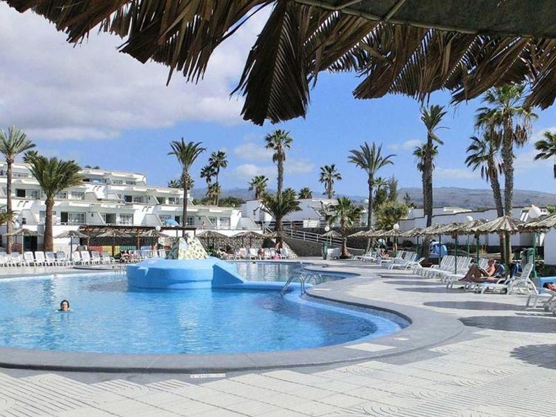 Großer Pool mit Liegestühlen, Palmen und weißen Gebäuden unter blauem Himmel.