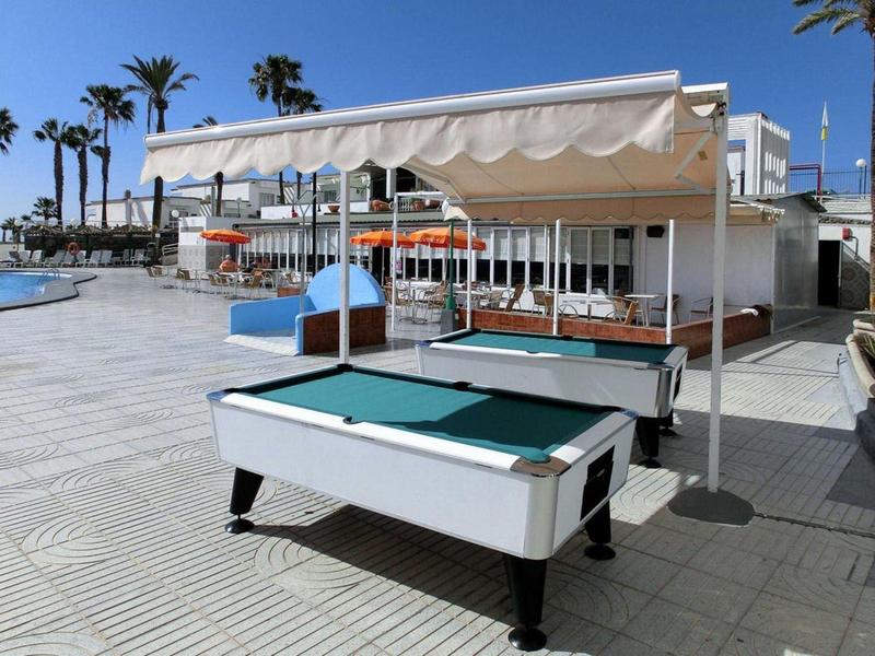 Ein luftiges Outdoorspiel mit Billardtisch unter Sonnenschutz, Palmen und klarem Himmel im Hintergrund.