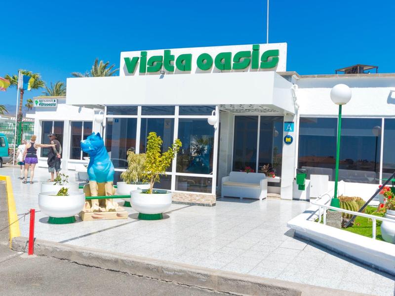 Helle Fassade eines Restaurants mit grünem Schriftzug "Vista Oasis" und Skulptur vor dem Eingang.