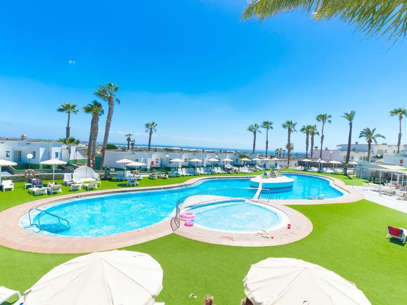 Großer Außenpool mit blauem Wasser, umgeben von Rasen, Palmen und weißen Sonnenschirmen.