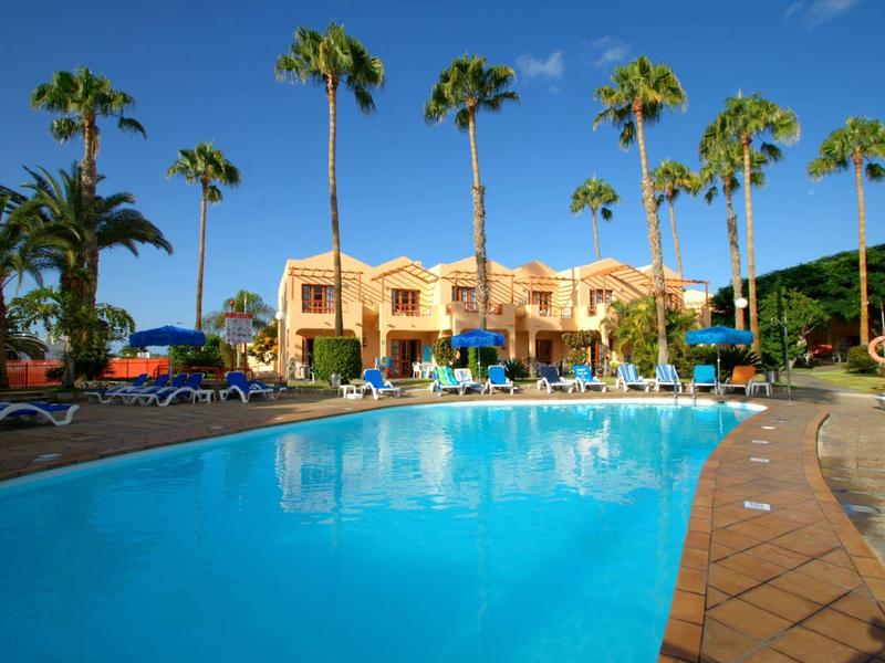Velký venkovní bazén před žlutými budovami s palmami pod jasnou modrou oblohou.