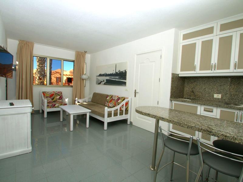 Světý obývací pokoj se šedou podlahou, pohovkou, stolem, TV a otevřenou kuchyní s barem.