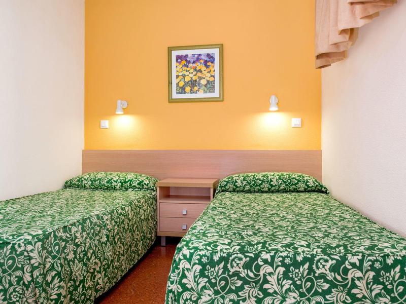 Hotelový pokoj se dvěma jednolůžky, zelenými vzorovanými přikrývkami a žlutou zdí s květinovým obrazem.