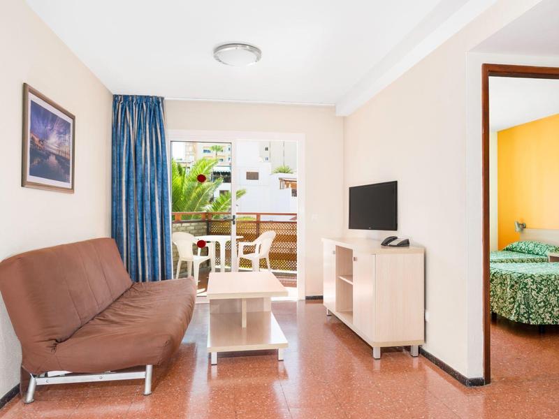 Světý obývací pokoj s hnědou pohovkou, TV, přístupem na balkon a výhledem do ložnice.