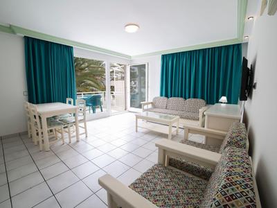 Helles Wohnzimmer mit weißen Möbeln, Fliesenboden und großen Fenstern mit blauen Vorhängen.