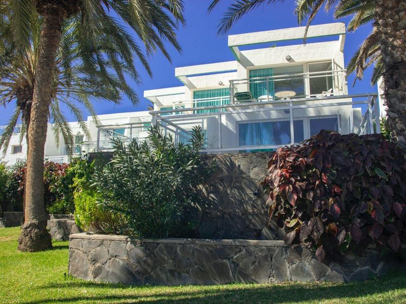 Modern white house with large glass fronts, surrounded by palm trees and green plants.