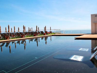 Schwarzer Pool mit Liegestühlen und Sonnenschirmen an der Küste bei klarem Himmel.