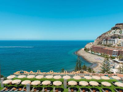 Blick auf Strand mit Sonnenschirmen, angrenzenden Hotels und blauem Meer unter klarem Himmel.