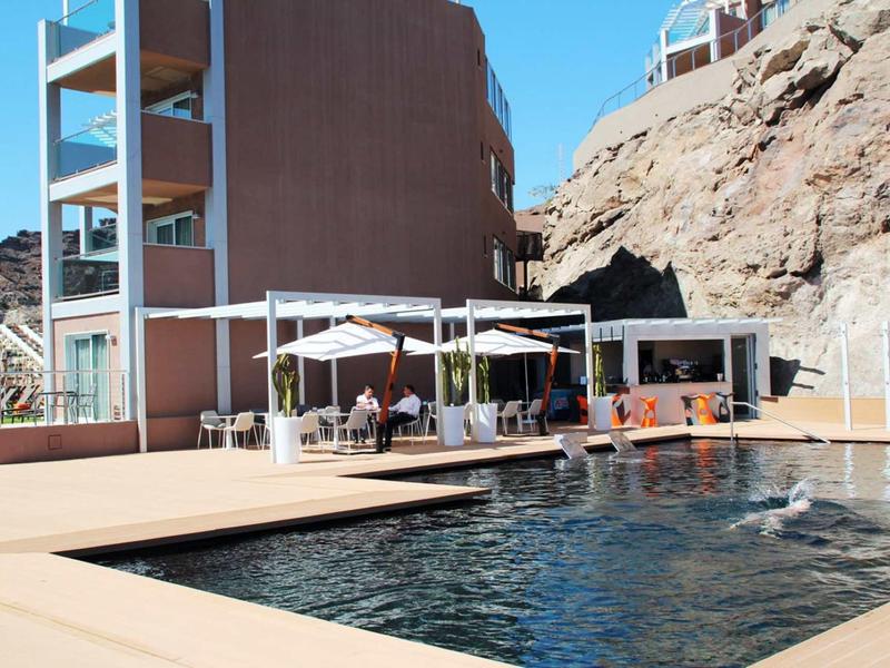 Moderner Pool mit Sonnenschirmen und Gästen vor Gebäuden und felsigem Hintergrund bei hellem Sonnenschein.