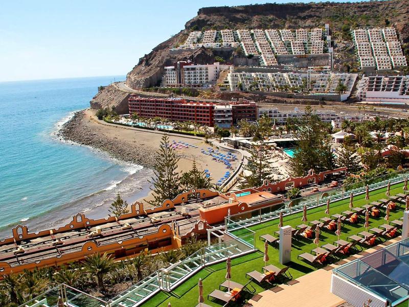 Küstenstadt mit Strand, Apartmenthäusern am Berghang und grünem Rasen mit Liegestühlen im Vordergrund.