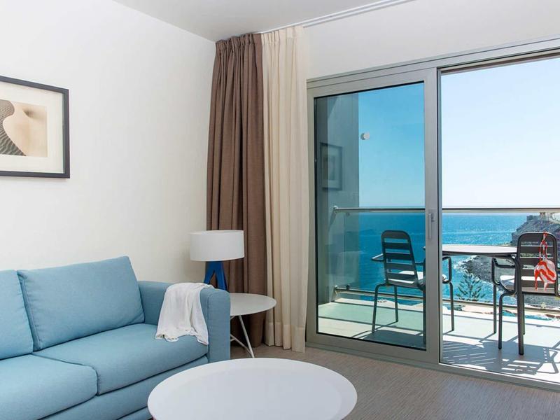 Helles Wohnzimmer mit blauem Sofa, weißem Tisch, Lampe und Blick auf Balkon und Meer.