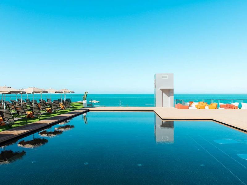 Moderner Infinity-Pool mit dunkelblauem Wasser, Sonnenliegen, Palmen und klarem blauem Himmel.