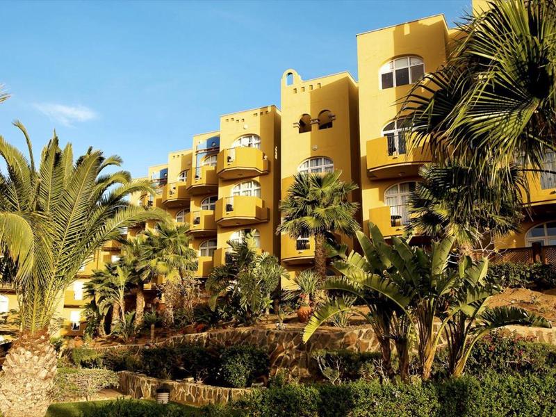 Gelbes Gebäude mit Balkonen, umgeben von Palmen und grünen Pflanzen unter blauem Himmel.