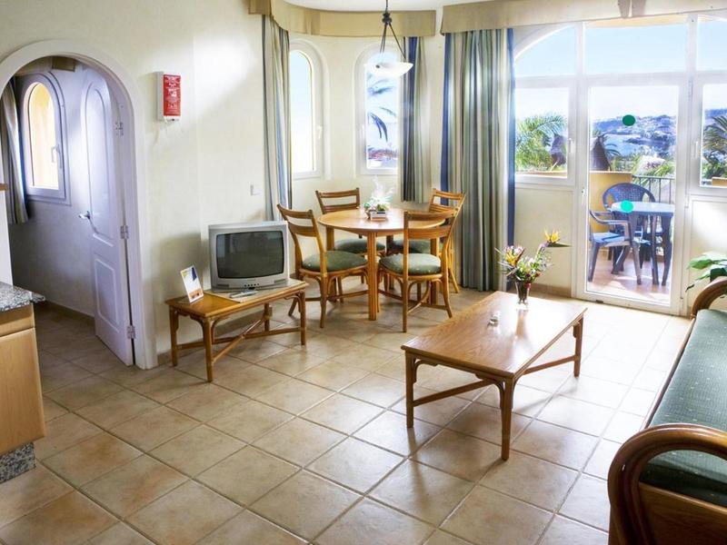 Helles Wohnzimmer mit Kachelfußboden, Holzmöbeln, Esstisch, Stühlen und Balkon mit Meerblick.