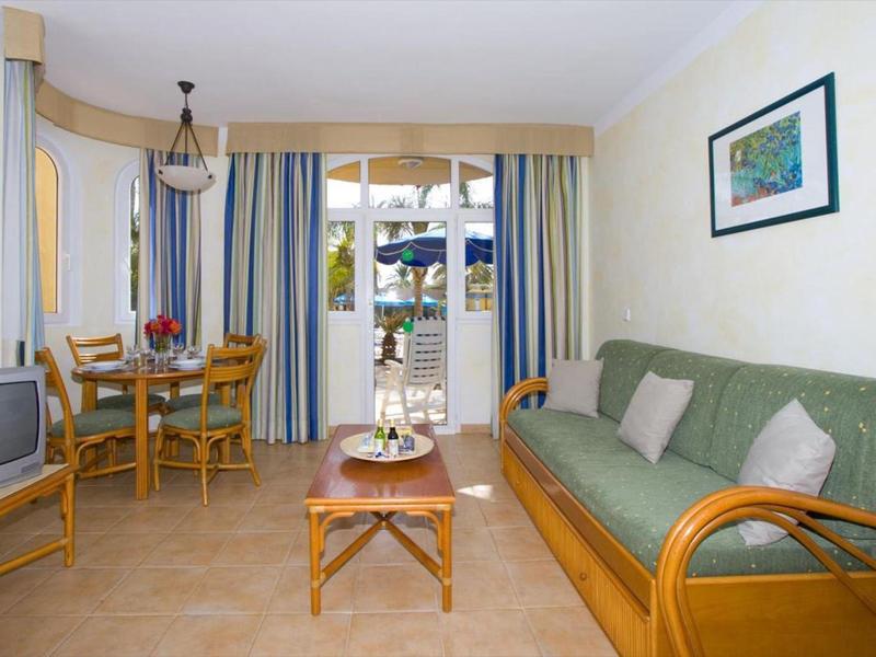 Helles Wohnzimmer mit grüner Couch, Holzmöbeln, kleinem Esstisch und Balkon mit blau-gelben Vorhängen.