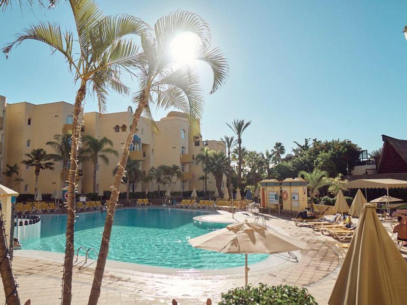 Ein sonniger Poolbereich mit Palmen, Sonnenschirmen und einem gelb-beigen Hotelgebäude im Hintergrund.