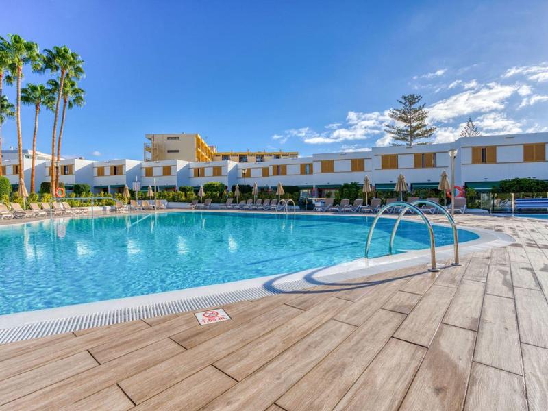 Hôtel avec grande piscine extérieure, palmiers et ciel dégagé en arrière-plan.