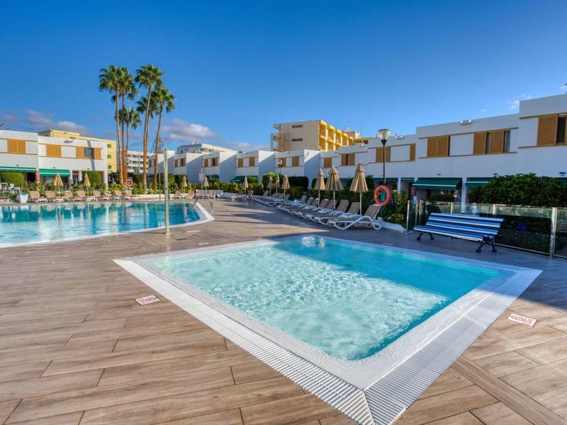 Espace piscine d'hôtel moderne avec petit bassin pour enfants, transats et bâtiments sous un ciel ensoleillé.