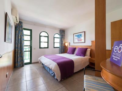 Chambre d'hôtel lumineuse avec lit double, accents violets et trois fenêtres en arc.