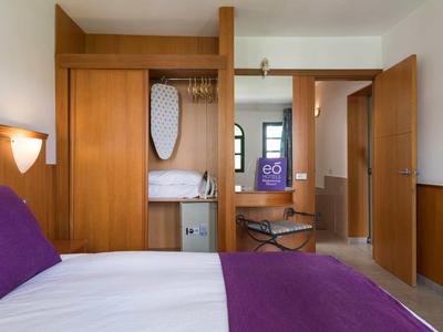 Chambre d'hôtel avec lit double, couvre-lit violet, meubles en bois et armoire ouverte.