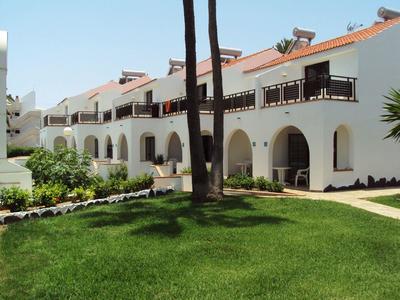 Weiße Reihenhäuser mit roten Dächern, Bögen und grünem Rasen im sonnigen Garten.