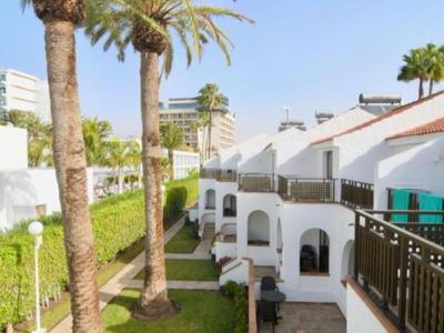 Weiße Reihenhäuser mit Balkonen und Palmen an sonnigem Tag neben grünem Rasen.