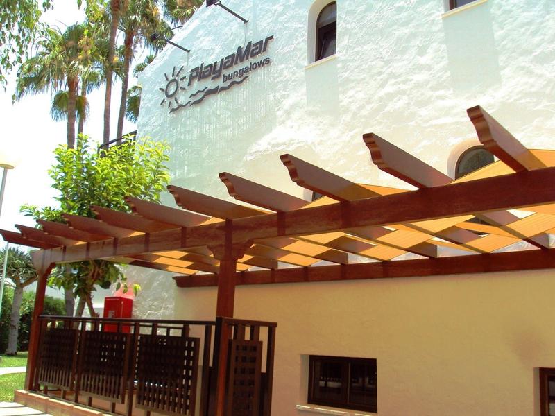 Exterior view of a hotel building with pergola and tropical trees in bright sunlight.